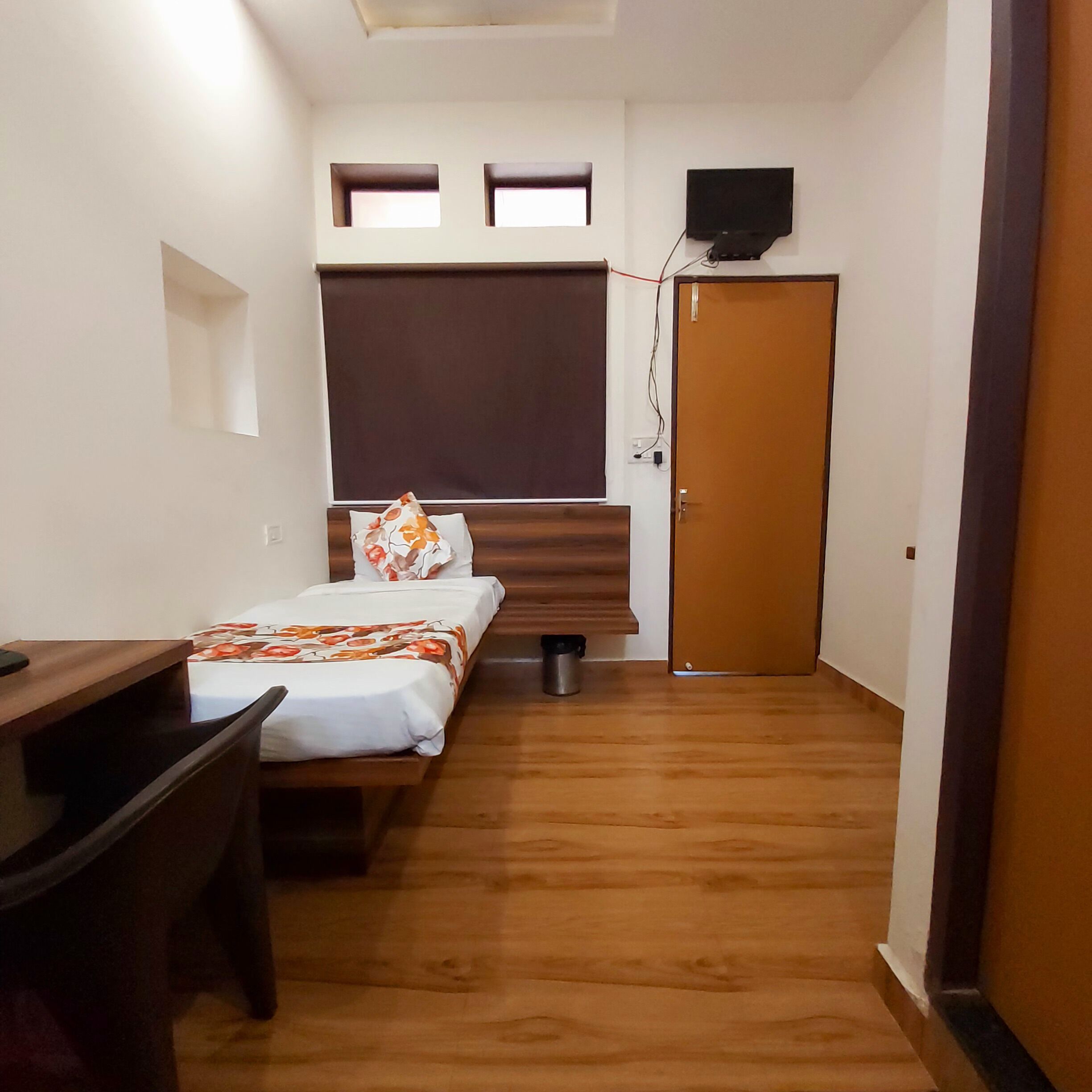 Una habitación pequeña y bien iluminada con una cama, un escritorio y un armario. Hay dos ventanas pequeñas y un televisor montado en la pared.