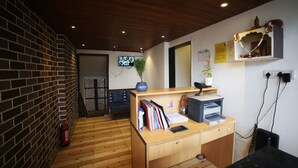 A reception desk with a wooden counter, a fire extinguisher, a plant, and a television screen in the background. - Hotel Park View (Bhopal)