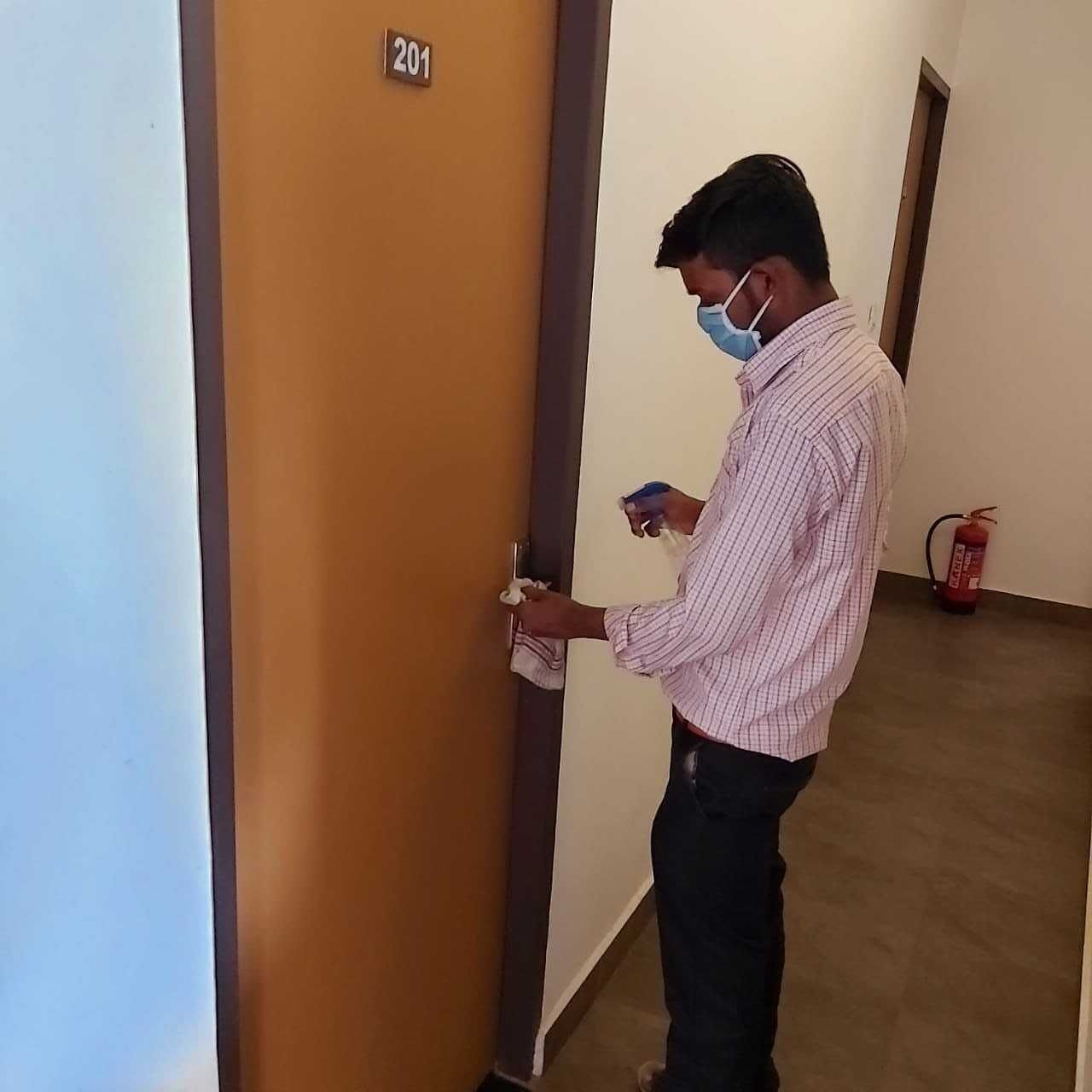 A person in a checkered shirt standing at a door with a number sign, holding a card and a cloth.