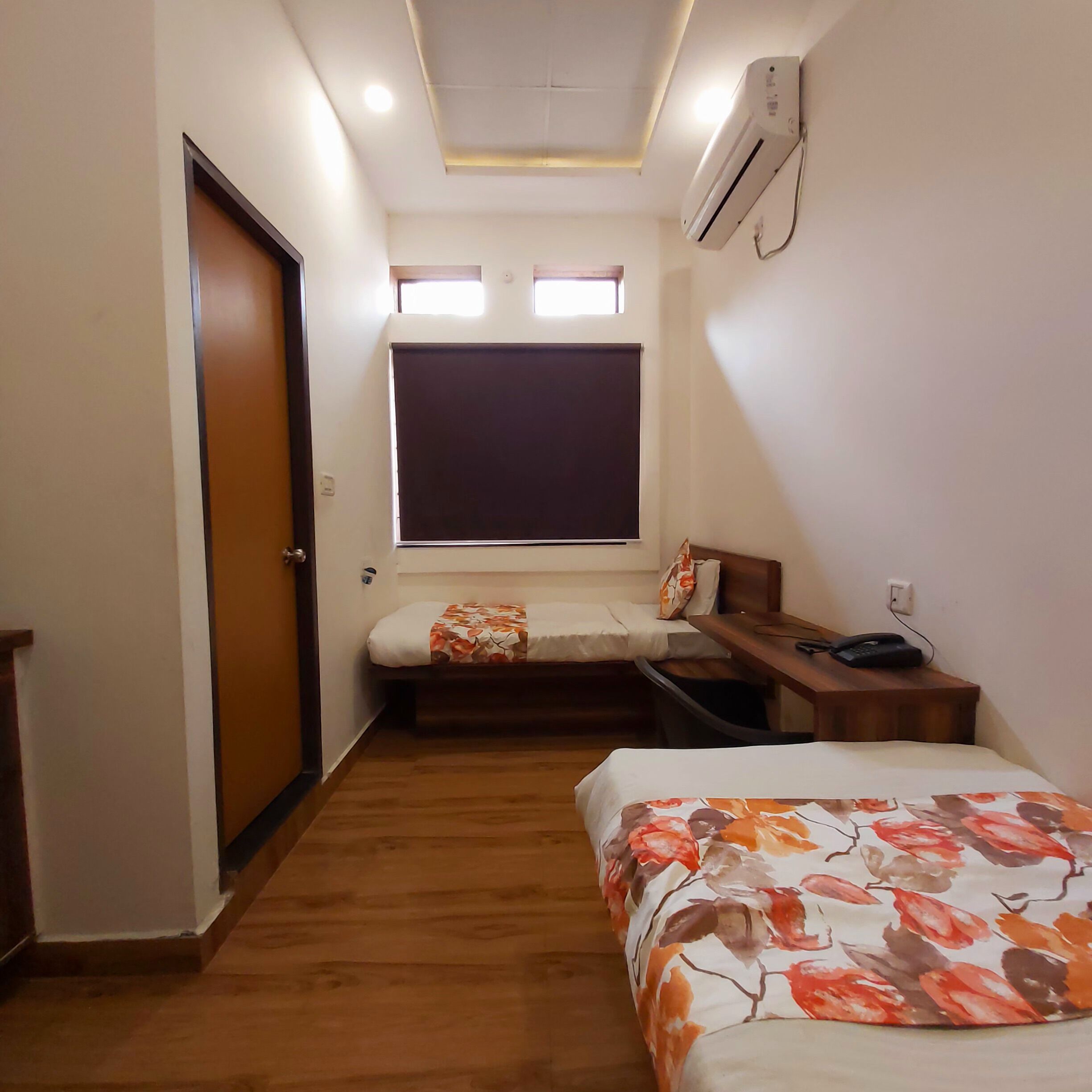 Habitación de hotel pequeña con cama, escritorio, silla, ventana con persianas y unidad de aire acondicionado.