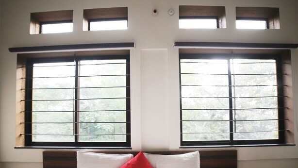 A bedroom with a large bed, two windows with blinds, a red pillow, and a white wall. - Hotel Park View (Bhopal)