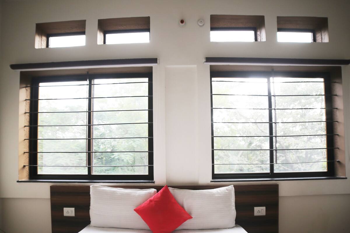 A bedroom with a large bed, two windows with blinds, a red pillow, and a white wall.