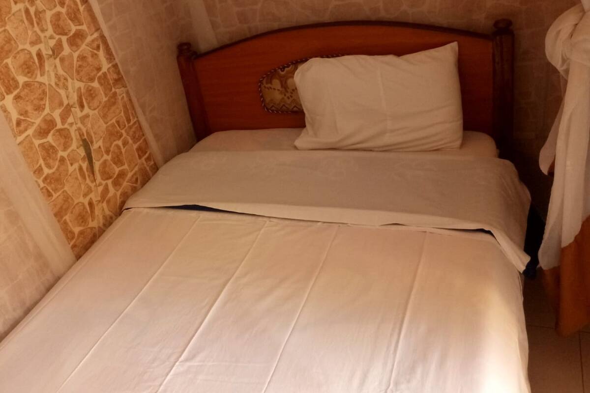 Un lit simple avec une tête de lit en bois, un oreiller blanc et une couvre-lit blanche, dans une pièce aux murs de pierre.