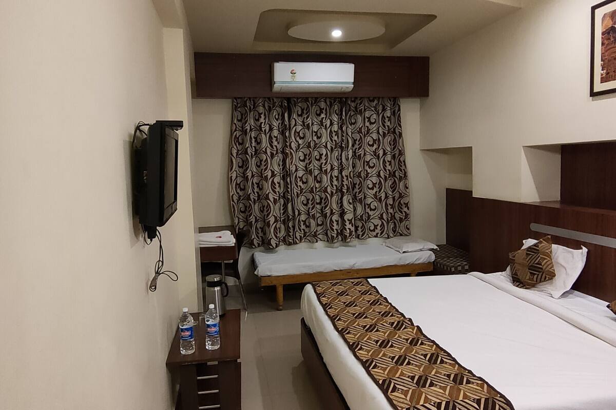 A hotel room with a bed, a desk, a chair, an air conditioning unit, and a wall-mounted TV.