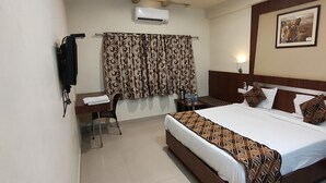 Habitación de hotel con cama, escritorio, silla, mesita de noche, aire acondicionado de pared, cortina y un cuadro en la pared.