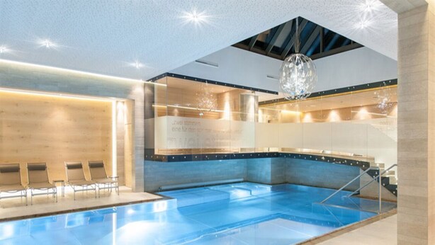 Piscina interior moderna com água cristalina, área de recepção em madeira e parede de vidro.