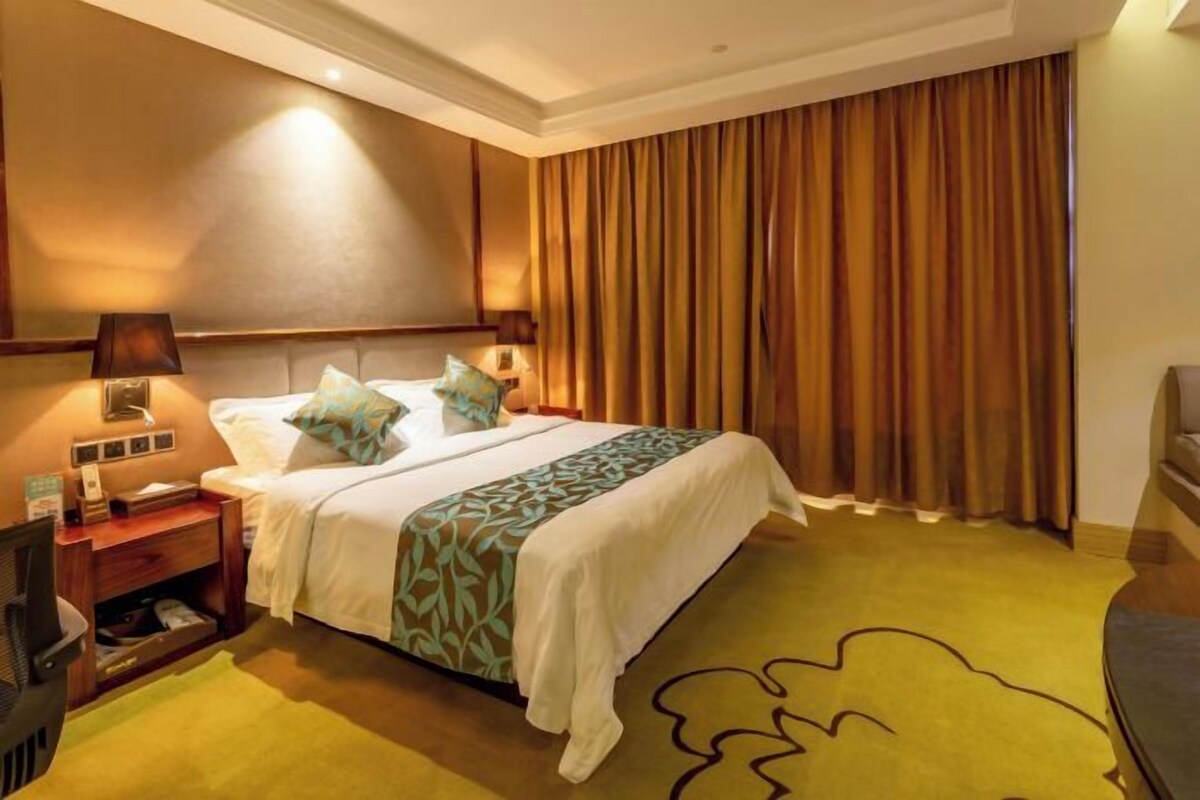 Habitación de hotel con cama, mesita de noche, lámpara, televisión y un amplio ventanal con cortinas.