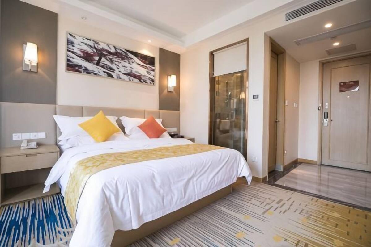 A hotel room with a bed, bedside tables, a wall-mounted lamp, a painting, a glass door, and a carpet.
