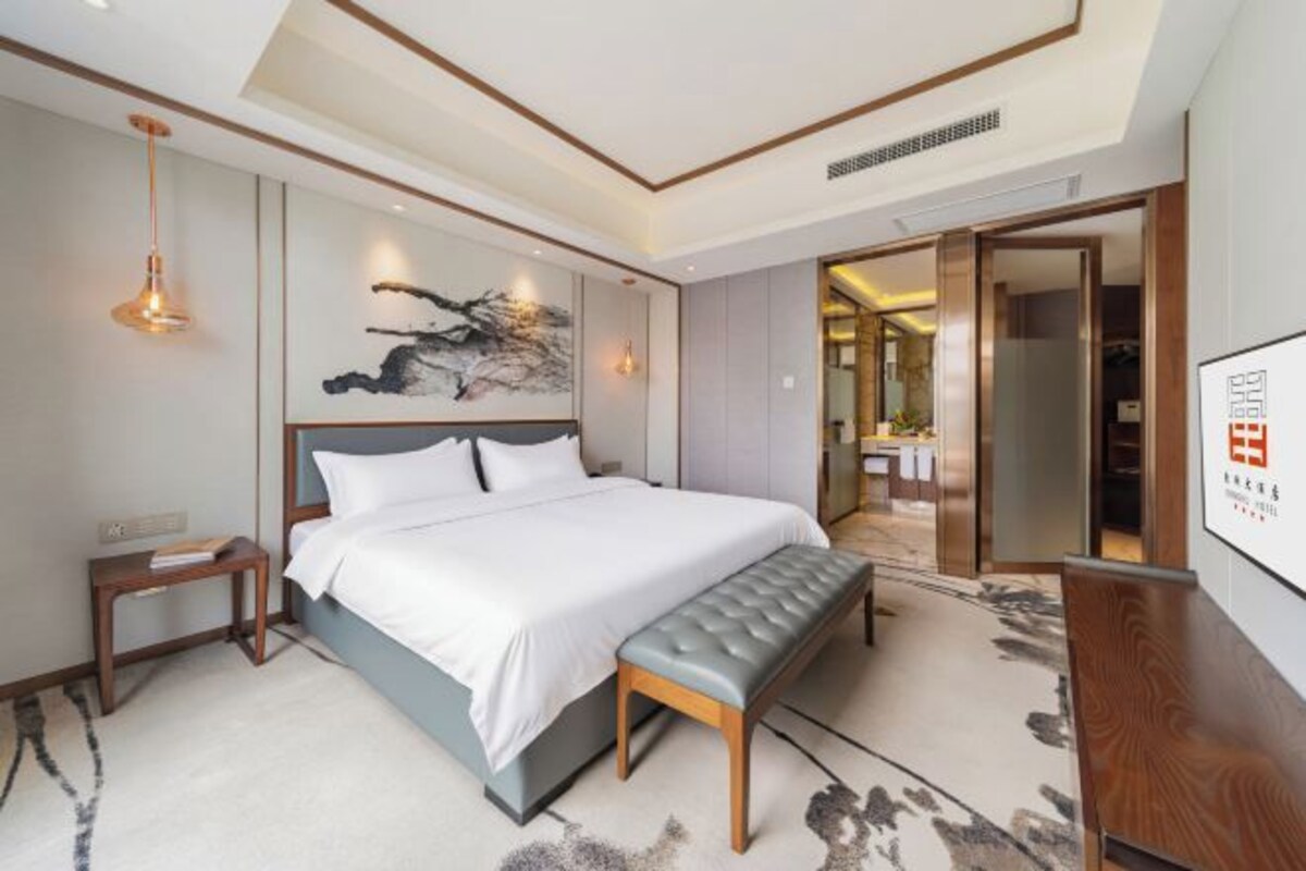 Quarto de hotel moderno com uma cama grande, um banquinho, uma mesa de cabeceira e uma TV fixada na parede.