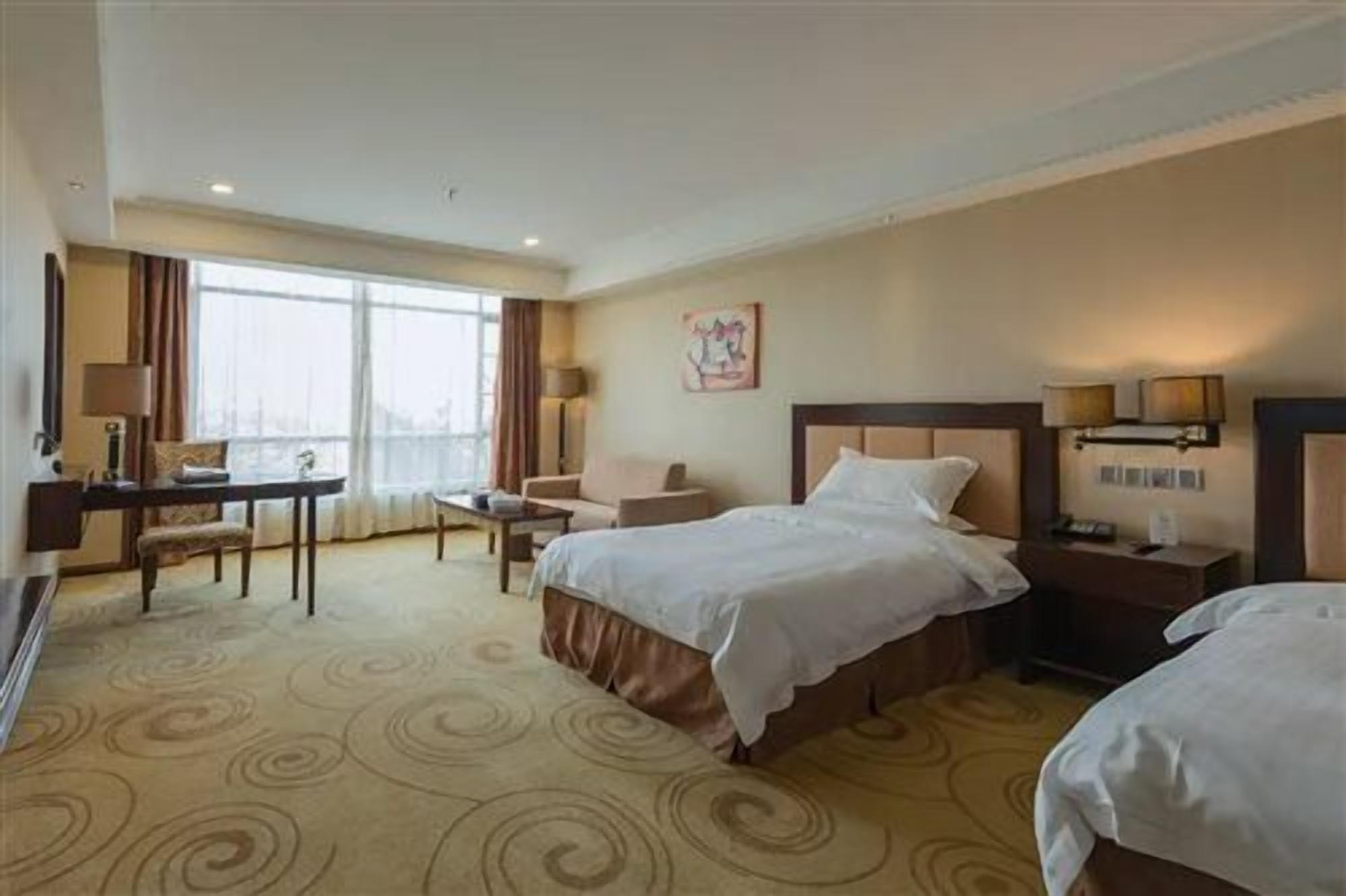 A hotel room with two beds, a desk, a chair, a lamp, and a window.