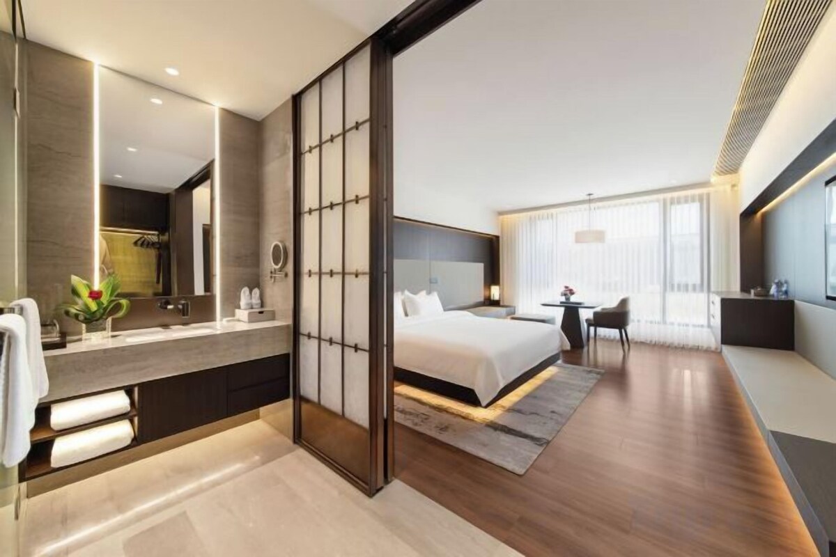 A modern hotel room with a large bed, a separate sitting area, a bathroom with a vanity, and a large mirror.