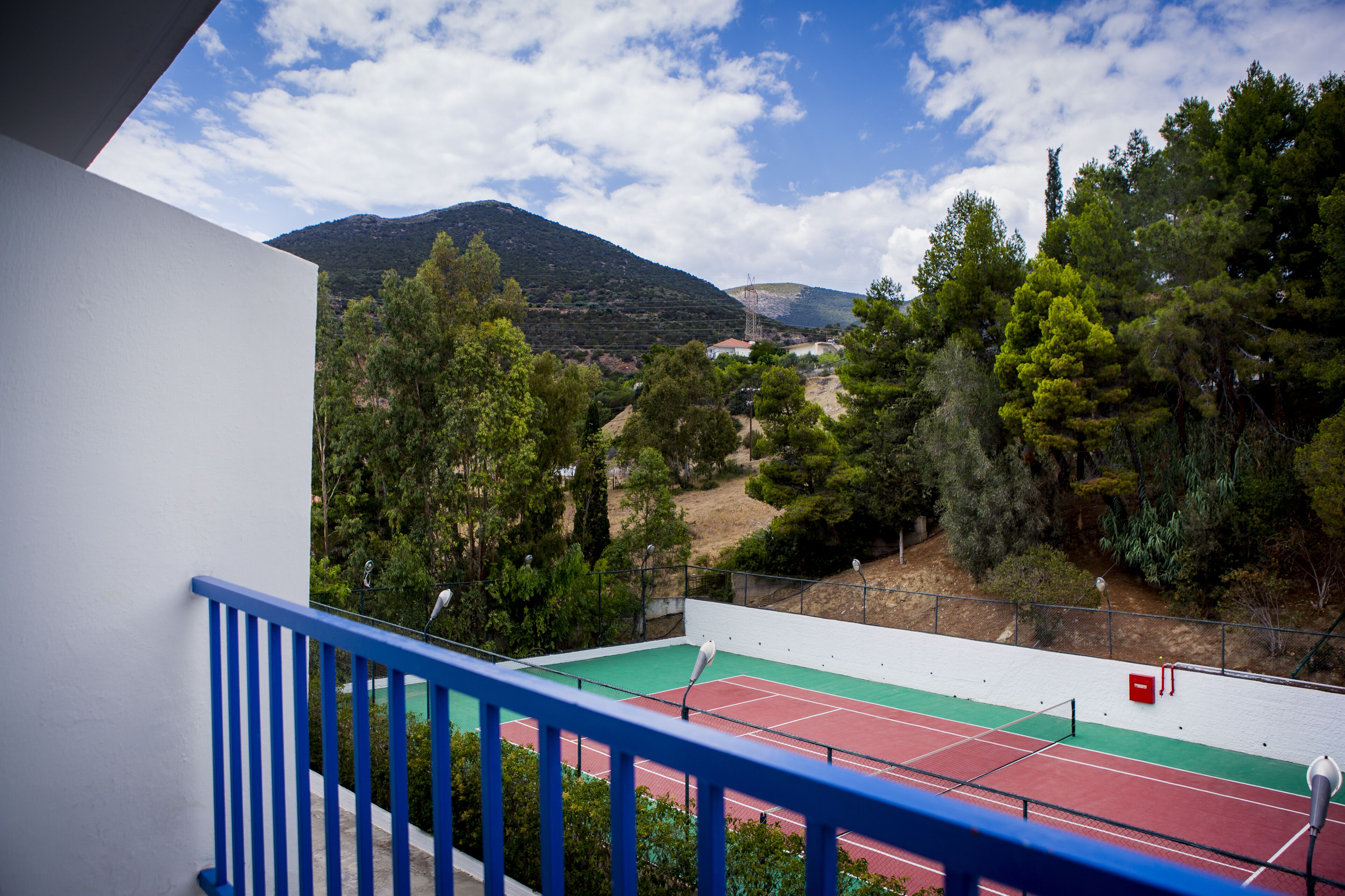 A tennis court with a red and green surface, surrounded by trees and a mountain in the background.