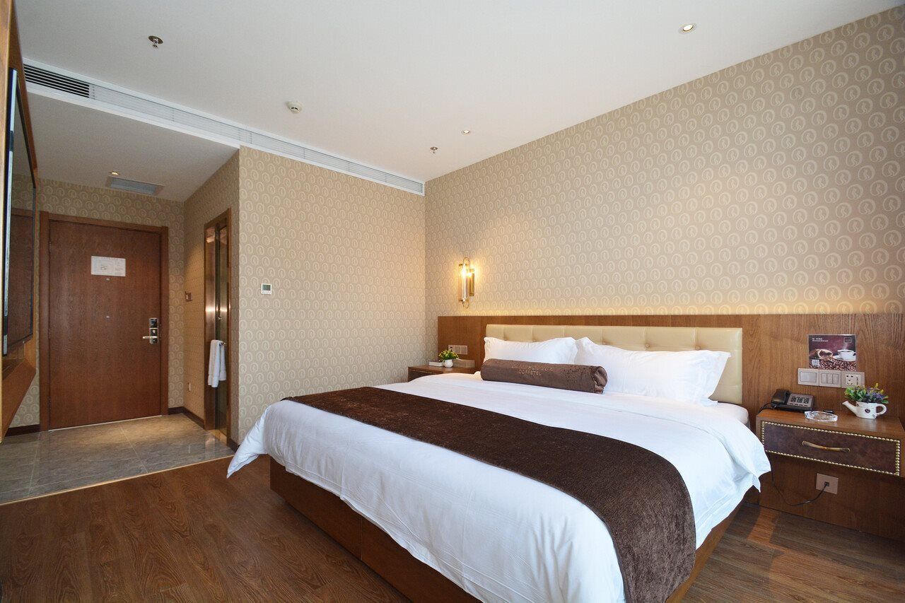 Habitación de hotel con una cama grande, piso de madera, cabecera con lámpara, mesita de noche con teléfono y una planta pequeña, y una puerta con un cartel.