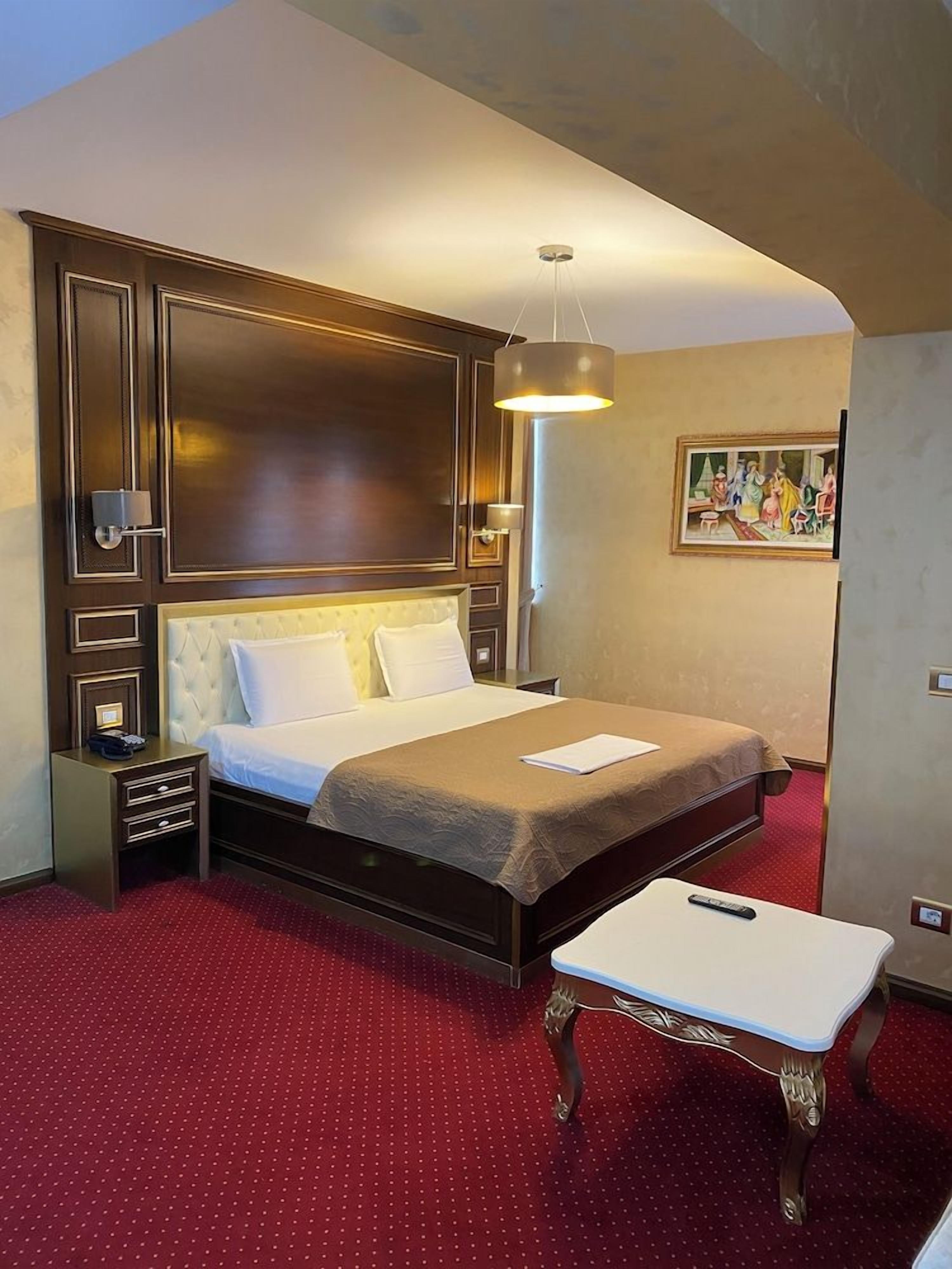 A hotel room with a large bed, a bedside table, a small table, and a painting on the wall.