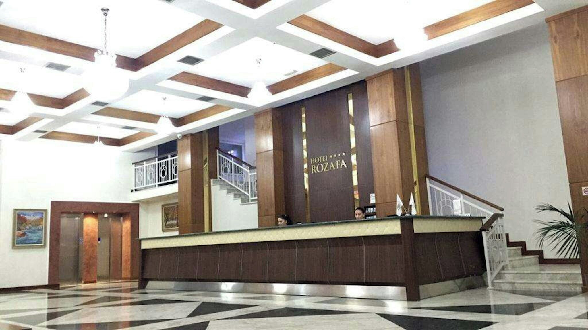 A hotel reception area with a modern design, featuring a large reception desk, a staircase, and a ceiling with geometric patterns.