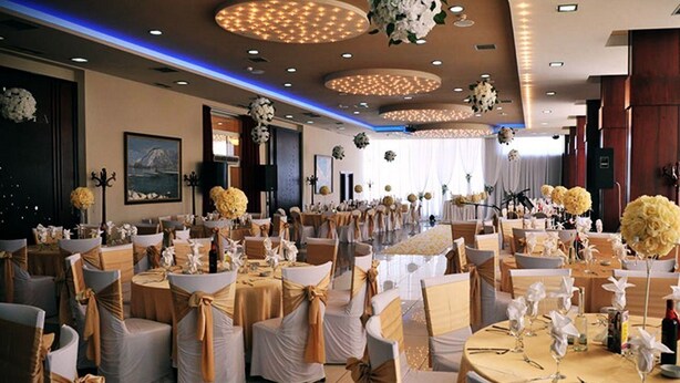 A banquet hall with round tables, chairs with gold sashes, and floral centerpieces. - Rozafa Hotel (Shkodër)