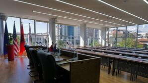 A conference room with multiple tables and chairs, flags of various countries, and large windows offering a view of buildings and greenery. - Rozafa Hotel (Shkodër)