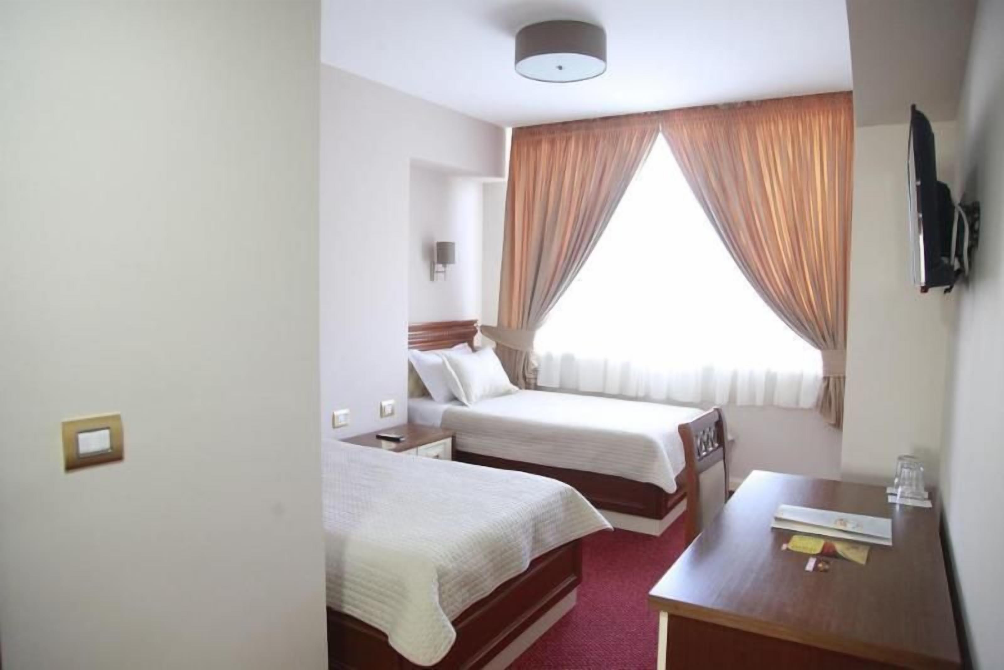 Habitación de hotel con dos camas, un escritorio, una silla, un televisor de pantalla plana montado en la pared y una ventana con cortinas.