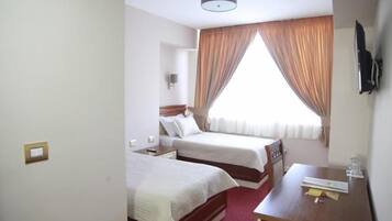 Habitación de hotel con dos camas, un escritorio, una silla, un televisor de pantalla plana montado en la pared y una ventana con cortinas.
