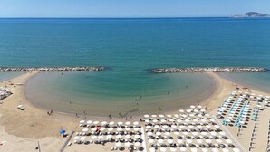 Una playa con un elemento acuático circular, una zona arenosa con sombrillas y sillas de playa, y un rompeolas rocoso.