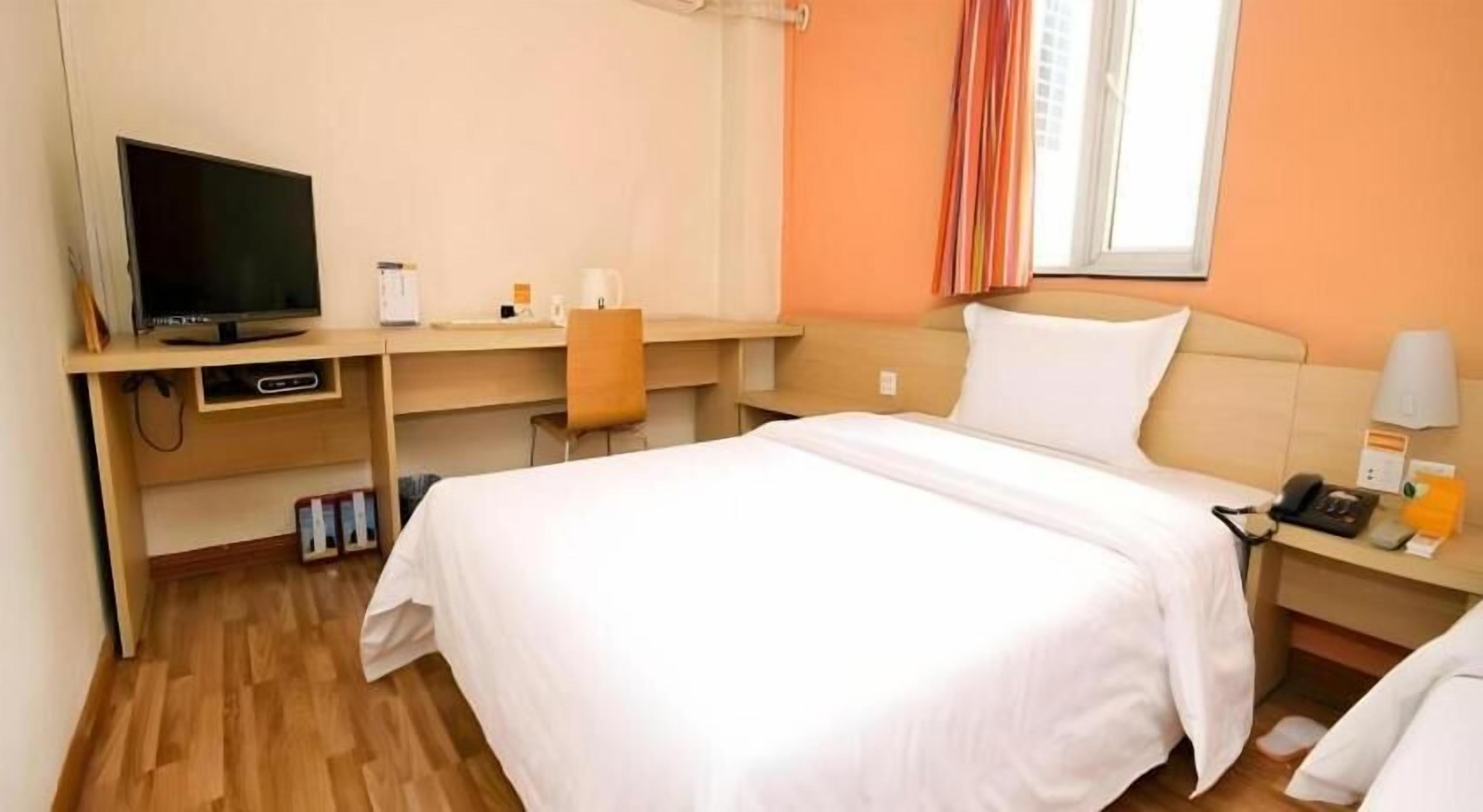 A hotel room with a bed, a desk with a computer, a TV, and a window with curtains.