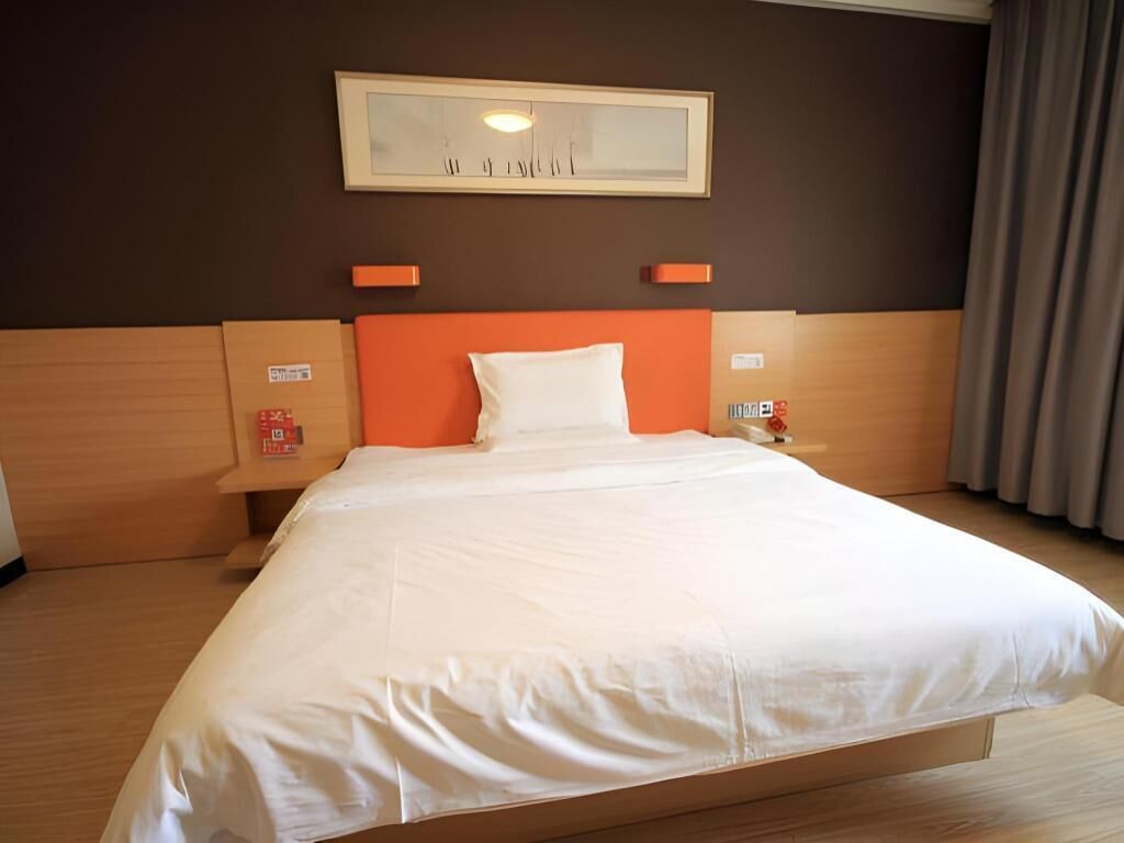A hotel room with a large bed, wooden headboard, white bedding, a framed artwork above the bed, and a nightstand with a phone and a red object.