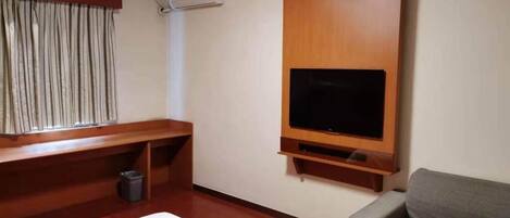 A hotel room with a bed, a TV mounted on the wall, a wooden bench, and a grey sofa.