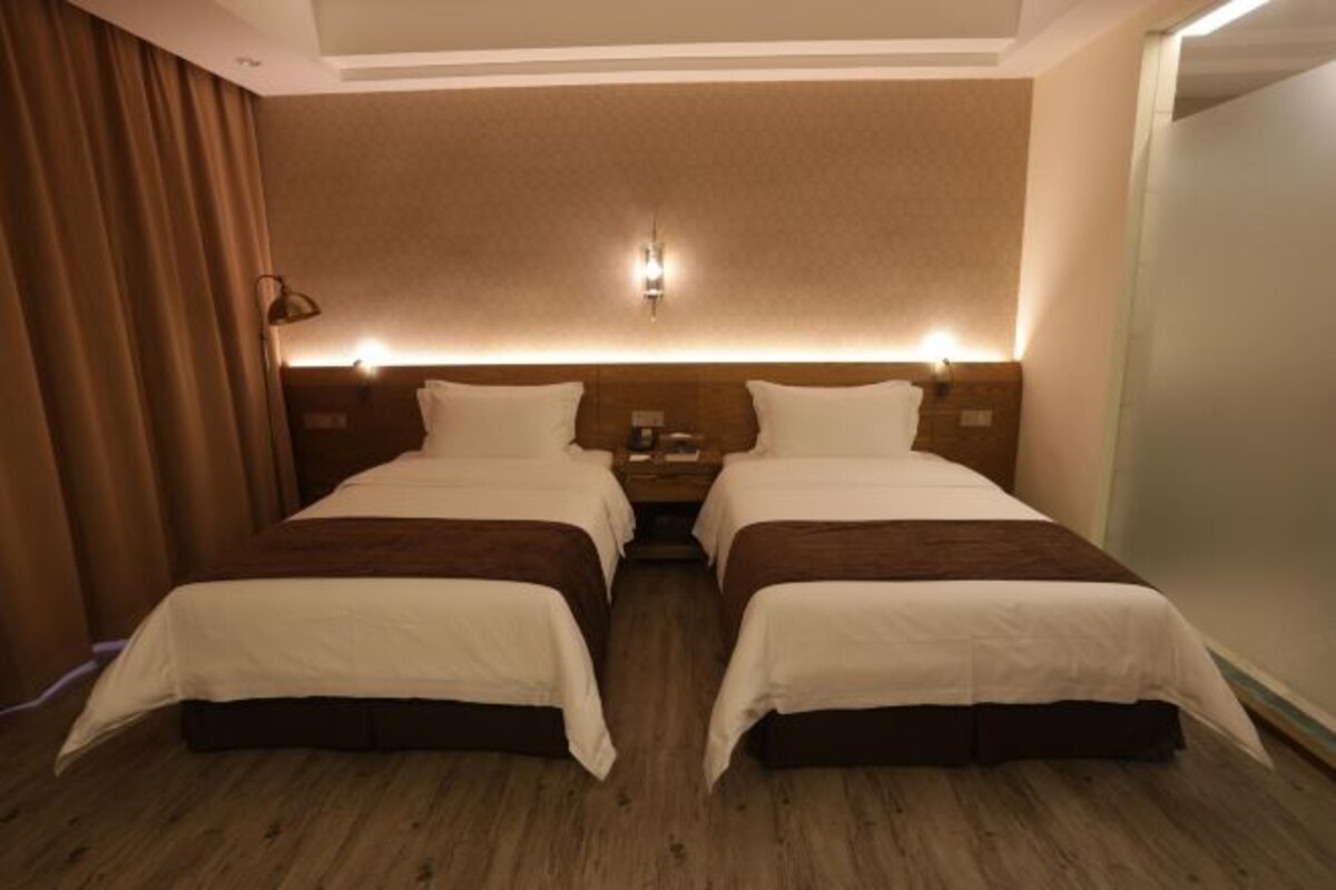 A double room with two beds, a wooden floor, a headboard with built-in lighting, and a curtain on the left.