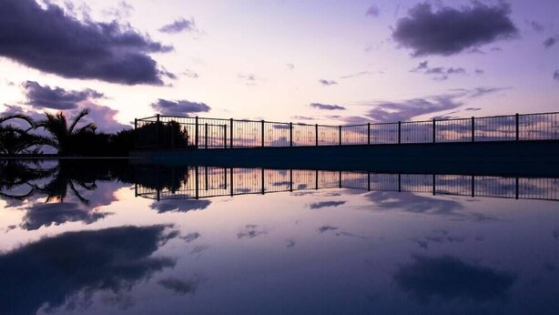 A bridge with a metal railing, reflecting in a calm body of water, with trees and a cloudy sky in the background. - Agriturismo Ninea (Ricadi)