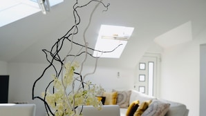 Una sala de estar moderna con un sofá blanco, una rama decorativa con flores blancas y un tragaluz arriba.