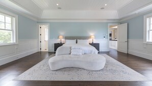 A spacious bedroom with a large bed, two bedside tables with lamps, a white bench, and a view of the outdoors through large windows. - Mansion Near the Bay. 7 bedrooms! Chef’s kitchen. (Southampton)