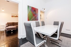 A modern dining area with a white table and grey chairs, a colorful abstract painting on the wall, and a sofa in the background.
