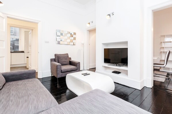 A modern living room with a grey sofa, a white coffee table, and a grey armchair. A flat-screen TV is mounted on the wall, and a decorative wall piece is visible.