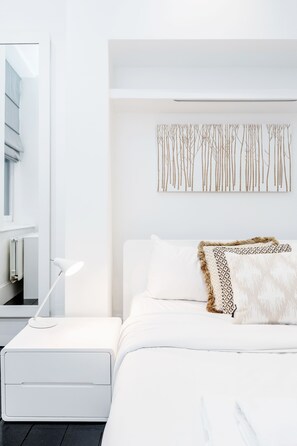 A modern bedroom with a white bed, a bedside table, a lamp, and a wall-mounted artwork.