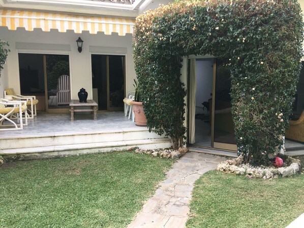 A covered patio area with outdoor seating, a potted plant, and a stone pathway leading to a door.