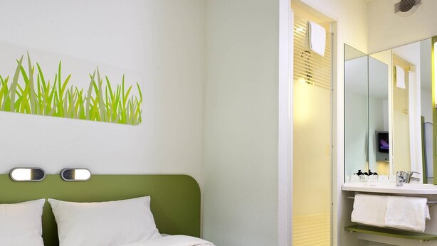 Een hotelkamer met een bed, een groen hoofdeinde, een wandlamp, een badkamer met spiegel, wastafel en handdoekrek.