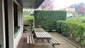 Een overdekt terras met een tafel en banken, een geplaveid pad en een haag op de achtergrond.