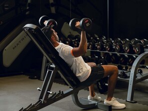 A person using a bench press with dumbbells, seated on a gym bench with a weight rack in the background. - Adagio Jeddah City Center (Jeddah)