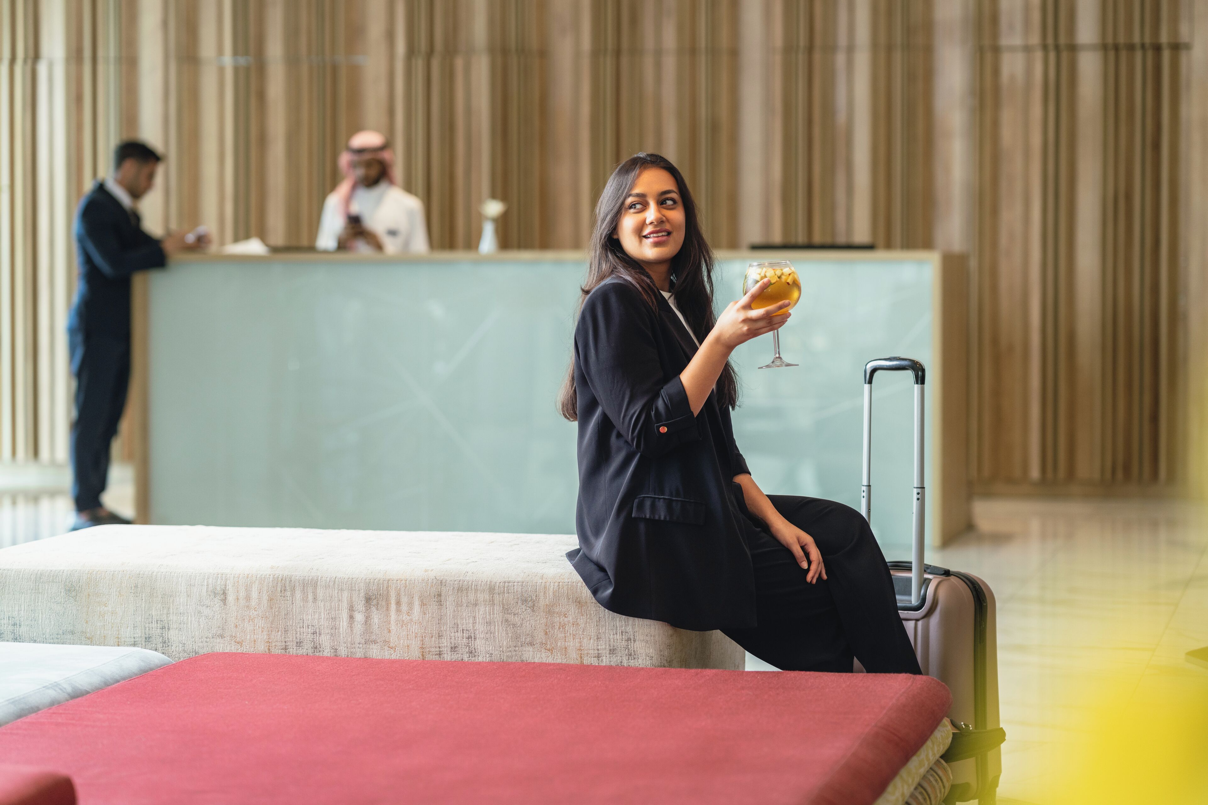 A woman sitting on a red couch in a hotel lobby with a suitcase and a drink.