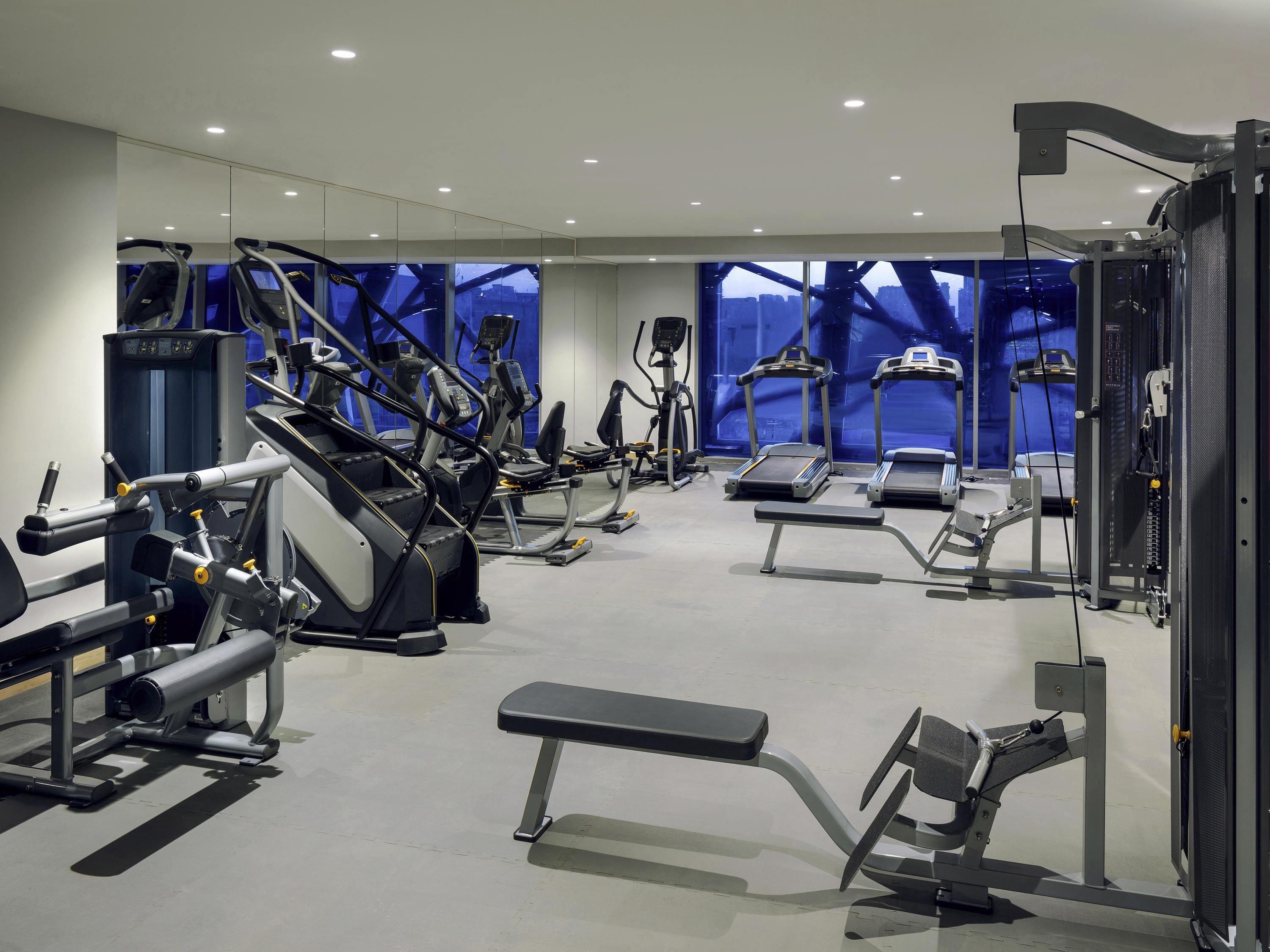 Un gimnasio con diferentes máquinas de ejercicio, una banca y ventanas amplias con vista a la ciudad.
