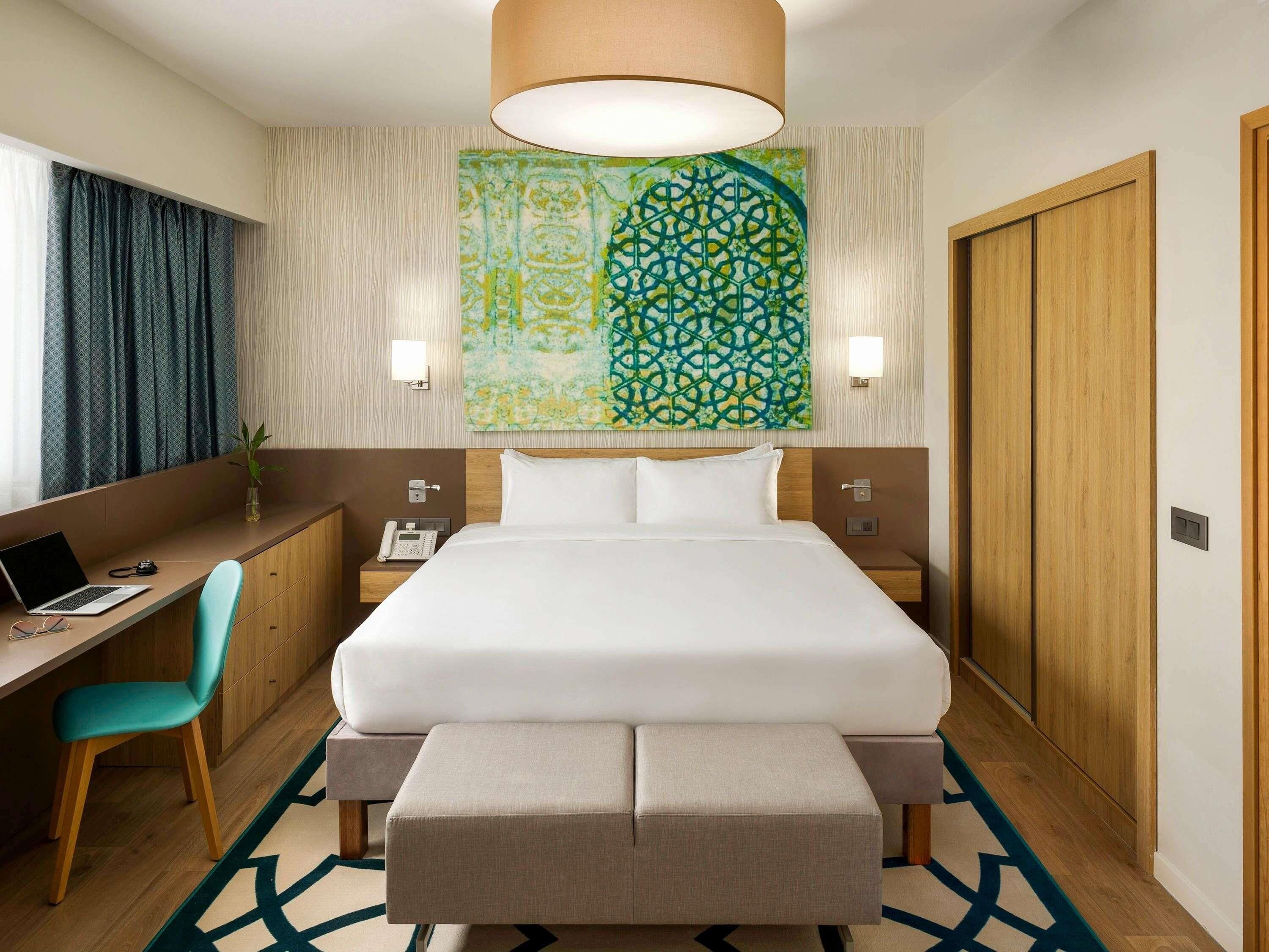 A hotel room with a bed, desk, chair, bedside tables, a lamp, and a wall art piece.