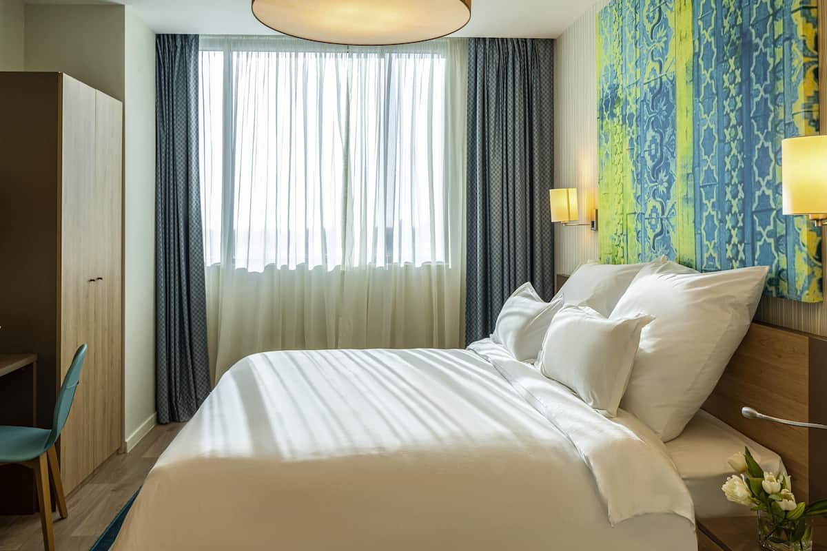 Una camera d'albergo con un letto, una sedia, un armadio, una finestra con tende e una parete decorata con un motivo.