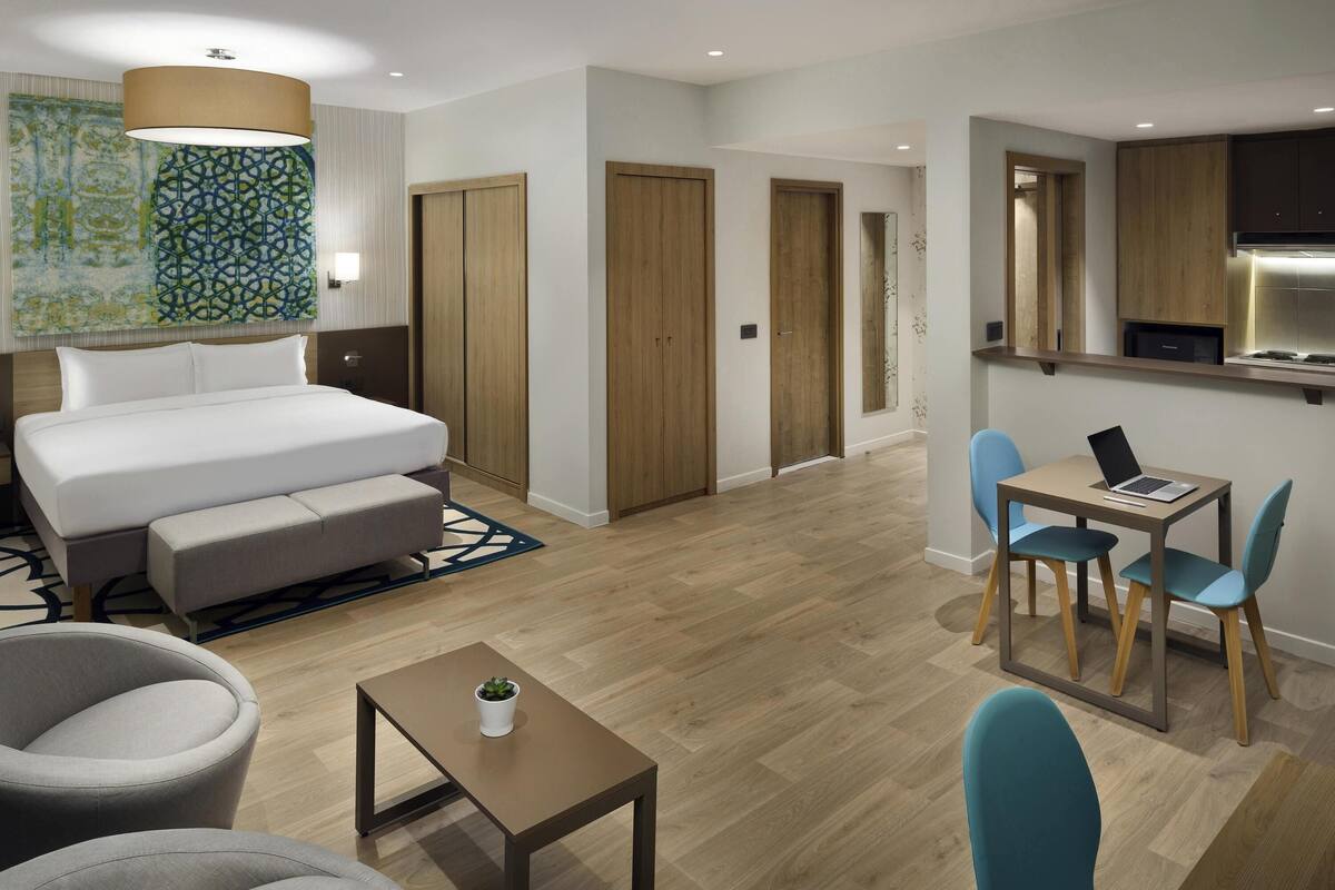 Una moderna camera d'albergo con un letto, un'area salotto, un piccolo tavolo, una scrivania con un portatile e un angolo cottura.