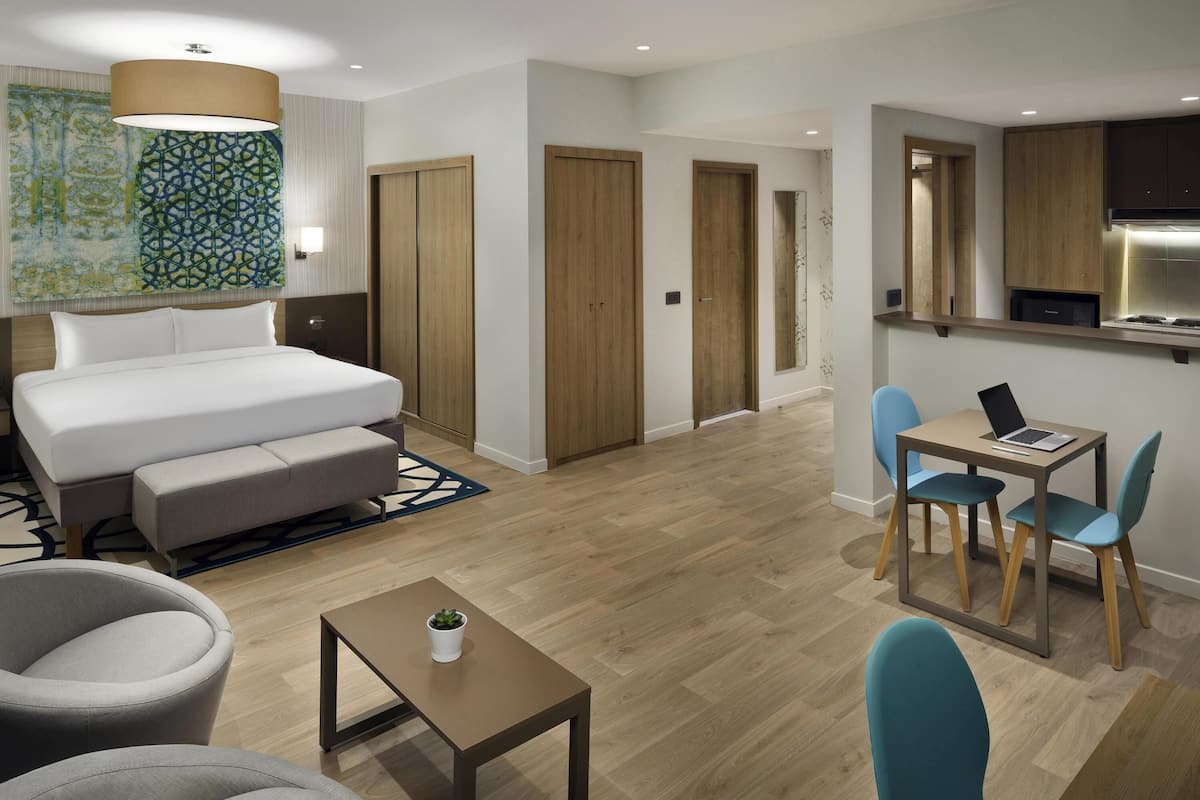 A modern hotel room with a bed, seating area, a small table, a desk with a laptop, and a kitchenette.