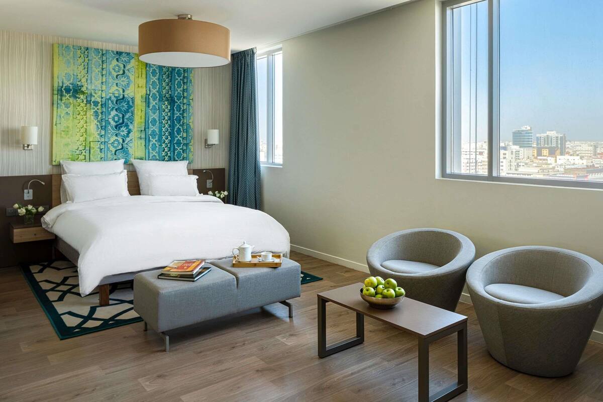 Camera d'albergo con un letto, due poltrone, un tavolino, vista sulla città e un'ampia finestra.