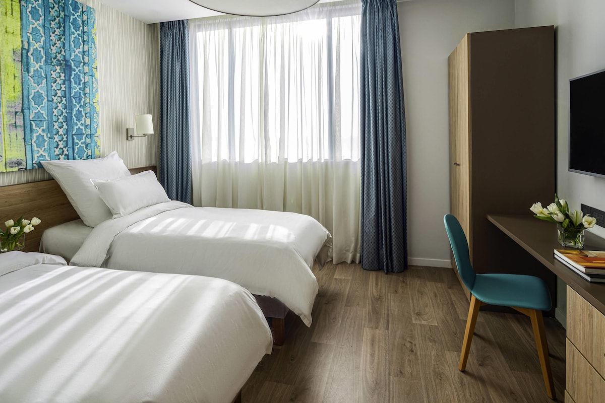 Habitación de hotel con dos camas, un escritorio, una silla, un televisor y una ventana con cortinas.