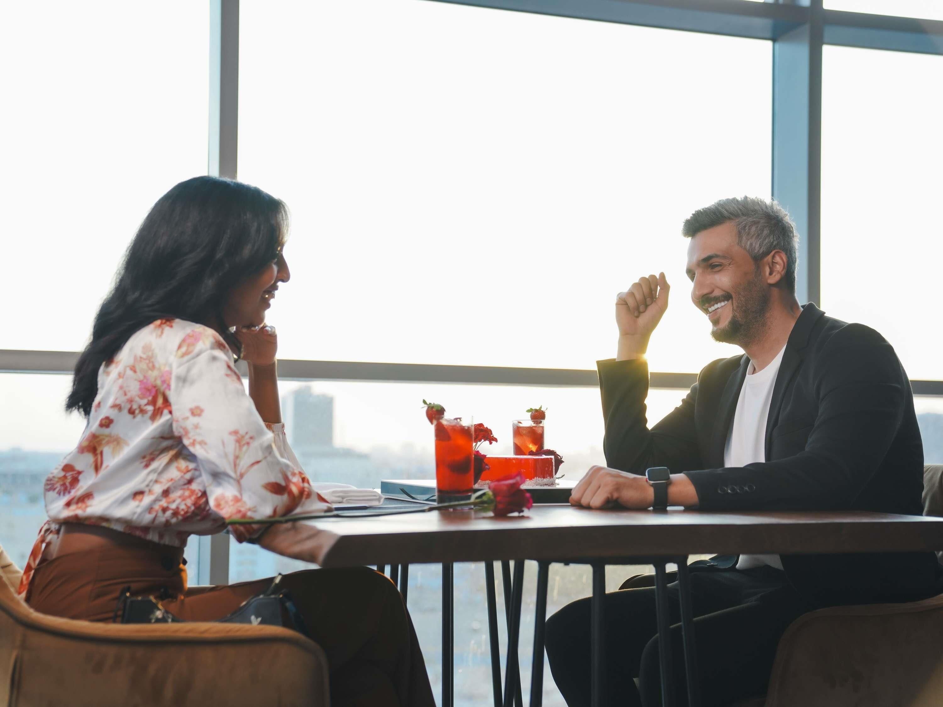 Twee personen zitten aan een tafel met drankjes, in een kamer met grote ramen en uitzicht.