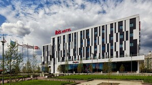 Een modern hotelgebouw met een opvallend ‘ibis HOTEL’ bord, een vlakke gevel met rechthoekige ramen, en een aangelegde voortuin met bomen en een pad.