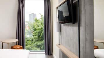 A modern hotel room with a large window, a flat-screen TV mounted on a wooden shelf, and a bed with a white headboard.
