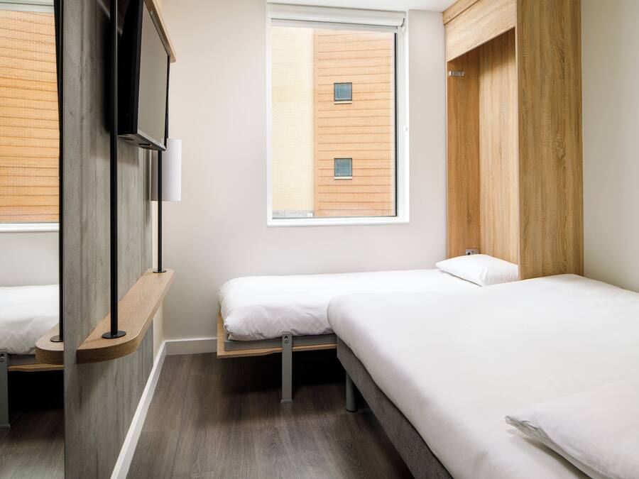 Una habitación de hotel moderna con dos camas individuales, un cabecero de madera, un ventanal grande y un televisor montado en la pared.